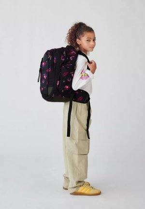 Sac à dos noir avec un motif floral coloré, doté de zippers et de bretelles réglables. Modèle portant un haut blanc et un pantalon cargo beige, tourné vers la gauche.