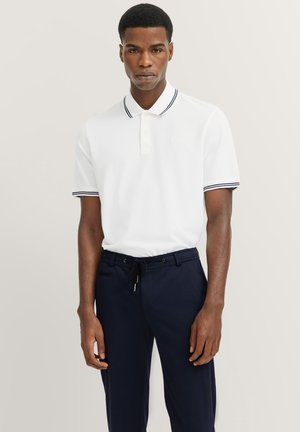 Weißes Poloshirt mit navyblauen gestreiften Akzenten am Kragen und an den Ärmeln, kombiniert mit dunkelblauen Hosen. Einfaches, klassisches Design.