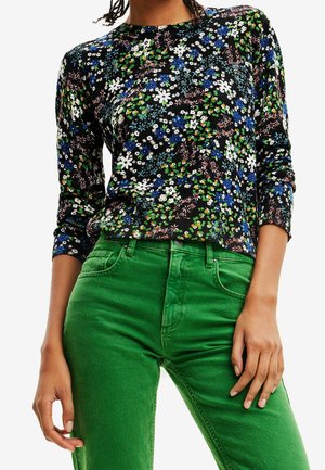 Kvinde iført en sort langærmet top med flerfarvet blomsterprint og lysegrønne bukser, med hænderne afslappet ned langs siden.