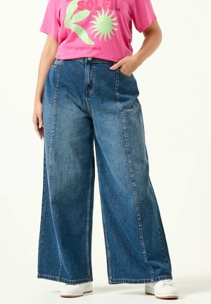 Person iført blå denimjeans med vide ben, hvide sneakers og en pink T-shirt med grøn solgrafik og rød tekst.