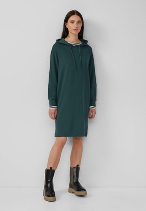Kapuzensweatkleid in dunkelblau mit langen Ärmeln, gestreiften Bündchen, einem lockeren Schnitt und seitlichen Schlitzen am Saum. Kombiniert mit Stiefeln.