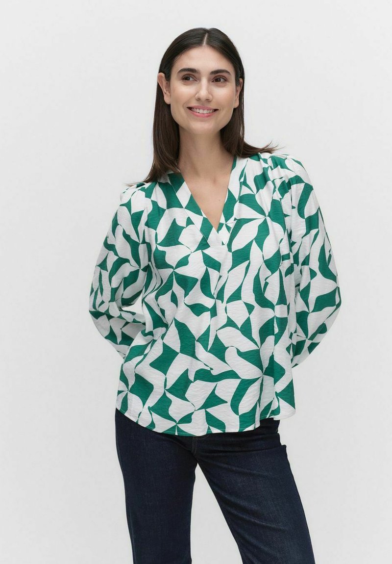 Grön och vit mönstrad blus med V-ringning, draperade ärmar och lös passform, tillsammans med mörka denimjeans. Slät tygstruktur.