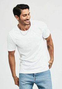 Vit kortärmad t-shirt i bomullstyg, rund halsringning, avslappnad passform, kombinerad med ljusblå denimjeans. Har tatueringar och ett silverhalsband.