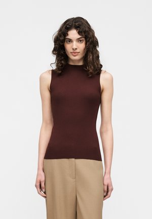 Femme aux cheveux bouclés foncés portant un haut marron sans manches côtelé et un pantalon beige, debout devant un fond clair uni.