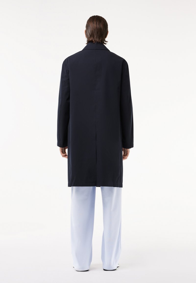 Abrigo largo de color navy con corte recto, abertura trasera y cuello ancho, llevado sobre pantalones azul claro. Textura suave; detalles mínimos.