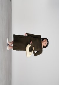 Μια γυναίκα φορώντας ένα μακρύ, σκούρο ελαιόχρωμο φόρεμα και λευκά μυτερά τακούνια, κρατώντας μια κρέμα τσάντα χειρός σε σχήμα ημισελήνου, στέκεται μπροστά σε ένα απλό φωτεινό φόντο.