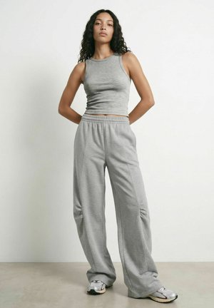 Femme debout, les mains derrière le dos, portant un crop top gris sans manches, un pantalon de jogging gris ample et des baskets argentées sur un fond uni.