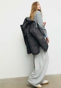 Maglione grigio e pantaloni di tuta grigi larghi, con un giubbotto imbottito nero appoggiato su una spalla e sneakers chiare ai piedi.