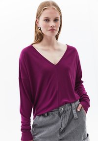 Pullover a maniche lunghe a V in vivace magenta, realizzato in un tessuto morbido. Abbinato a pantaloni grigi testurizzati con dettaglio di bottone e cinturino.