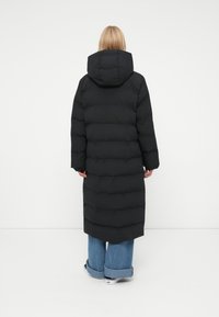 Abrigo de plumas negro con una silueta larga y capucha, que presenta un patrón acolchado horizontal. Combinado con jeans de pierna ancha y zapatos casuales.