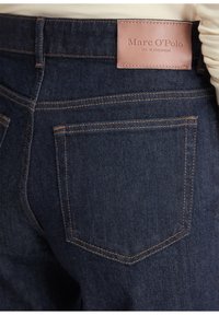 Dunkelblaue Jeans aus Denim verfügen über ein braunes Lederetikett mit der Aufschrift "Marc O’Polo" am Taillenbund, kontrastierenden orangefarbenen Nähten und Gesäßtaschen.