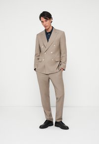 Beige διπλο-κουμπωτό κοστούμι με ασημένια κουμπιά, κομμένο για τέλεια εφαρμογή, υφασμάτινη υφή, συνδυασμένο με μπλε πουκάμισο και μαύρα παπούτσια, σε φόντο λευκό.