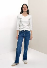 Femme debout portant une chemise blanche à manches longues, un jean bleu et des baskets blanches sur un fond clair uni.