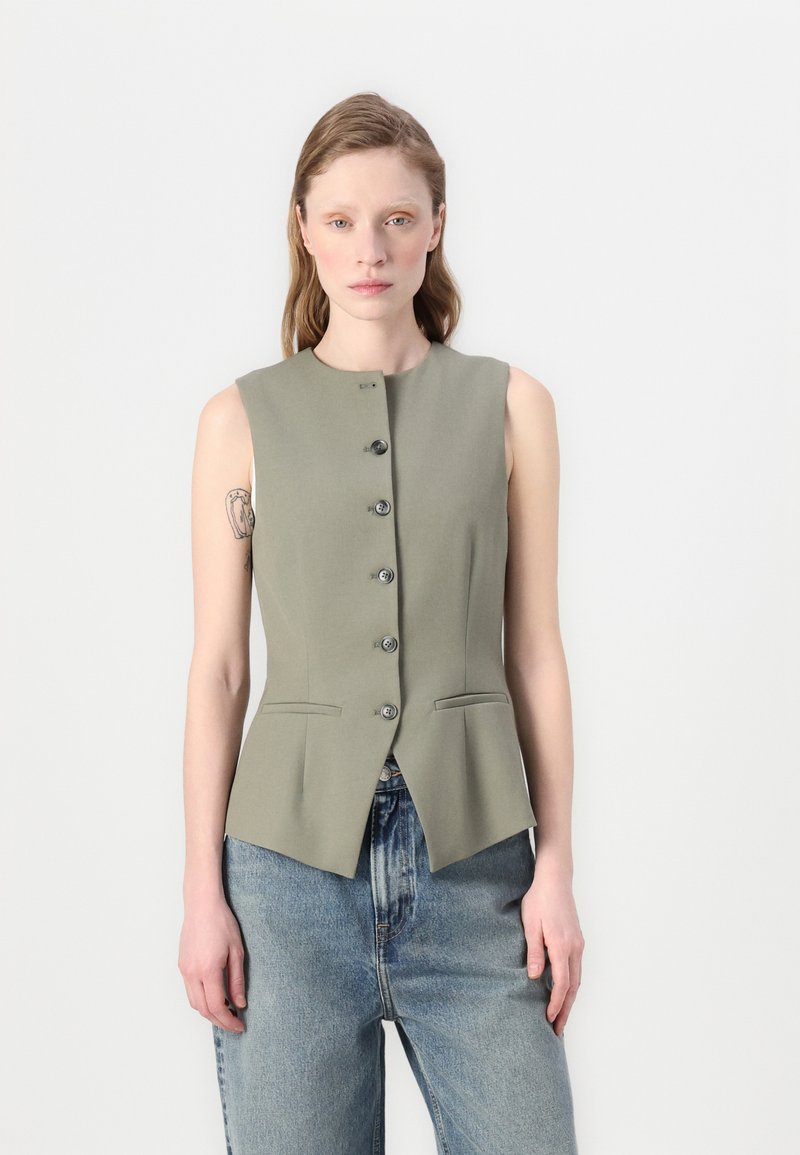 Ermeløs grønn vest laget av glatt materiale, med høy hals, seks knapper foran og to lommer. Fitted design med utvidet skjortekant.