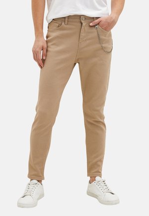 Uomo che indossa pantaloni beige slim-fit con una catena d'argento e sneakers bianche, in piedi contro uno sfondo semplice, mano in tasca.