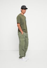 Oliven grøn t-shirt med grafik, cargo-bukser i matchende farve, hvide sneakers og en sort kasket med logo foran, i stående stilling.