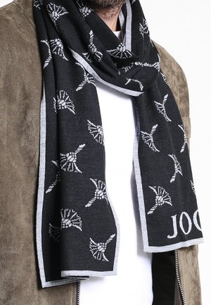 Écharpe noire avec motifs floraux blancs et bords gris portée sur une veste en daim marron et une chemise blanche, couvrant partiellement le cou.