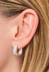 Primo piano di un orecchio sinistro con capelli biondi che indossa un orecchino a cerchio quadrato in argento.