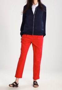 Marinblå zip-up tröja med ribbad nederkant, matchad med ljus röda byxor och svarta sandaler med remmar, med en slät yta och skräddarsydd passform.