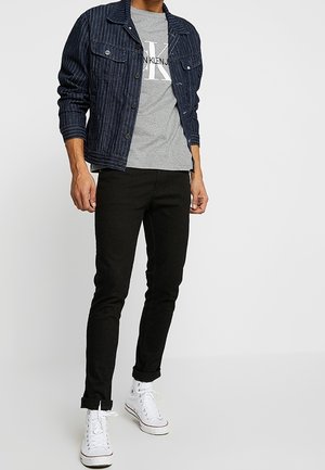 Giacca di jeans con righe verticali, t-shirt grigia con grande logo, pantaloni neri aderenti e sneakers bianche.