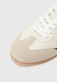 Beige sneaker met bovenwerk van suède en synthetisch materiaal, ronde neus, witte veters en een zool van gumrubber. Opvallende naden en kleurcontrasten.