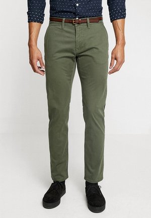 Homme portant un pantalon slim vert olive avec ceinture marron, une chemise bleu marine à boutons avec des pois blancs, et des chaussures noires à lacets.