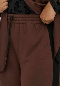 Ruke podešavaju tamnosmeđe elastične hlače s crnom šnuricom, uparene s crnom majicom s cvjetnim uzorkom.