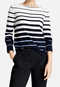 Gestreifter langarm Pullover in Weiß, Navy und Hellblau. Weicher Strickstoff mit einem Bootsausschnitt. Kombiniert mit schwarzen Hosen.