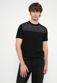 T-shirt preta de manga curta com um painel texturizado em padrão preto no peito, decote redondo e um corte regular. Combinada com jeans escuros.