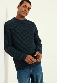 Ein Mann trägt einen dunkelblauen gerippten Pullover und blaue Jeans, lehnt an einer weißen Wand und hat die Hände locker vor sich gefaltet.