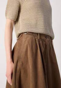 Haut beige à manches courtes en maille, rentré dans une jupe en velours côtelé marron taille haute avec passants de ceinture et plis, visible du nombril à la main.