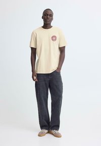 Beige katoenen T-shirt met een rond borstlogo, gecombineerd met zwarte loszittende jeans en grijze sneakers met een gestructureerd ontwerp.