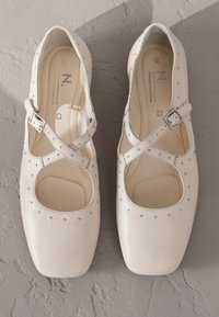 Fehér bőr balettcipő lekerekített négyzet alakú orral, keresztpántokkal és apró, fémes szegecsekkel az élén. Simogató textúra.