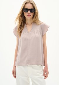 Blusa di un pallido mauve con scollatura a V, pieghe e maniche a sbuffo. Abbinata a pantaloni bianchi, indossata da un modello con occhiali da sole su uno sfondo neutro.