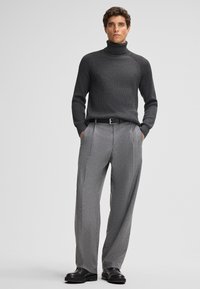 Grauer gerippter Rollkragenpullover, kombiniert mit hellgrauen, gefalteten Hosen. Ein schwarzer Gürtel und glatte schwarze Schuhe runden das Outfit ab.