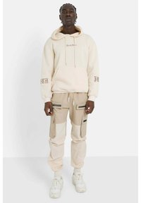 Felpa beige con tasca frontale, testo "Sixth June" sulle maniche. Pantaloni cargo beige con pannelli a contrasto e tasche con zip. Sneakers bianche.