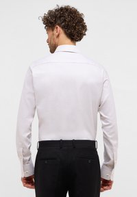 Camicia col colletto di un beige chiaro con tessuto testurizzato, maniche lunghe e polsini con bottoni, abbinata a pantaloni neri con tasche posteriori e bottoni.