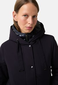 Czarna kurtka zewnętrzna z błyszczącym, izolowanym kapturem. Posiada gładką tkaninę, regulowane troczki i metalowe napy.