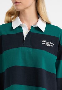 Maglietta da rugby a righe verdi e blu navy con colletto bianco, caratterizzata da un logo sul lato sinistro del petto, realizzata in tessuto misto cotone.