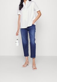 Vit skjorta med krage, blå korta jeans och vita remsandaler. Ett hållaraccessoar i handen ger en elegant, minimalistisk touch.