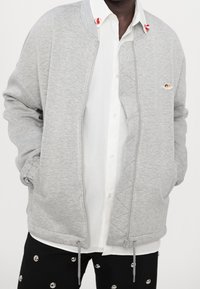 Grijs gewatteerd sweatshirt met geribbelde manchetten en kraag, voorzien van een ritssluiting aan de voorkant en twee zijzakken. Afgewerkt met een klein logodetail.