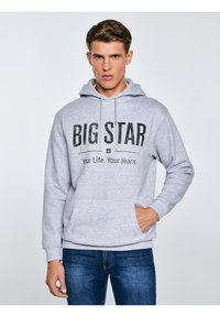 Noor mees, kes kannab halli kapuutsiga pusa, millel on tekst "BIG STAR Your Life. Your Jeans.", siniste teksadega, seisab valge tausta ees.