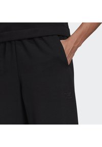 Handen vilar i fickan på svarta Adidas sweatpants med elastisk midja och tonal Adidas-logotyp på låret.