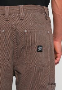 Pantaloni a righe marroni e bianche realizzati in tessuto testurizzato, dotati di tasche posteriori e di un'etichetta nera con logo sul lato destro.