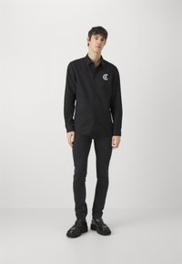 Black button-up shirt with a logo on the left chest, paired with dark slim-fit trousers and black lace-up shoes. Simple, modern design.