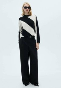 Pull en tricot avec des rayures diagonales noires et crème, associé à un pantalon noir taille haute et des talons noirs élégants. Lunettes de soleil noires incluses.