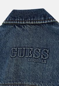 Tył kurtki dżinsowej z kołnierzem i tłoczonym logo "GUESS ORIGINALS" wyszywanym niebieską nicią w odcieniu dopasowanym do tkaniny.