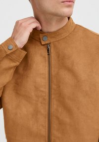 Veste en suède marron avec col montant, bouton-pression argenté au cou, fermeture éclair et texture douce. Gros plan sur la partie supérieure du corps.