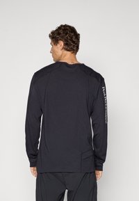 Mann, der sich abwendet, trägt ein schwarzes langärmliges Shirt mit dem Schriftzug "Peak Performance" am rechten Ärmel und schwarze Hosen.