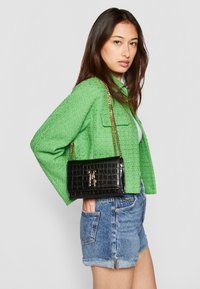 Svart faux krokodilleveske med gullkjedehank, rektangulær form og distinkt teksturmønster. Modellen har på seg en grønn jakke og denimshorts.